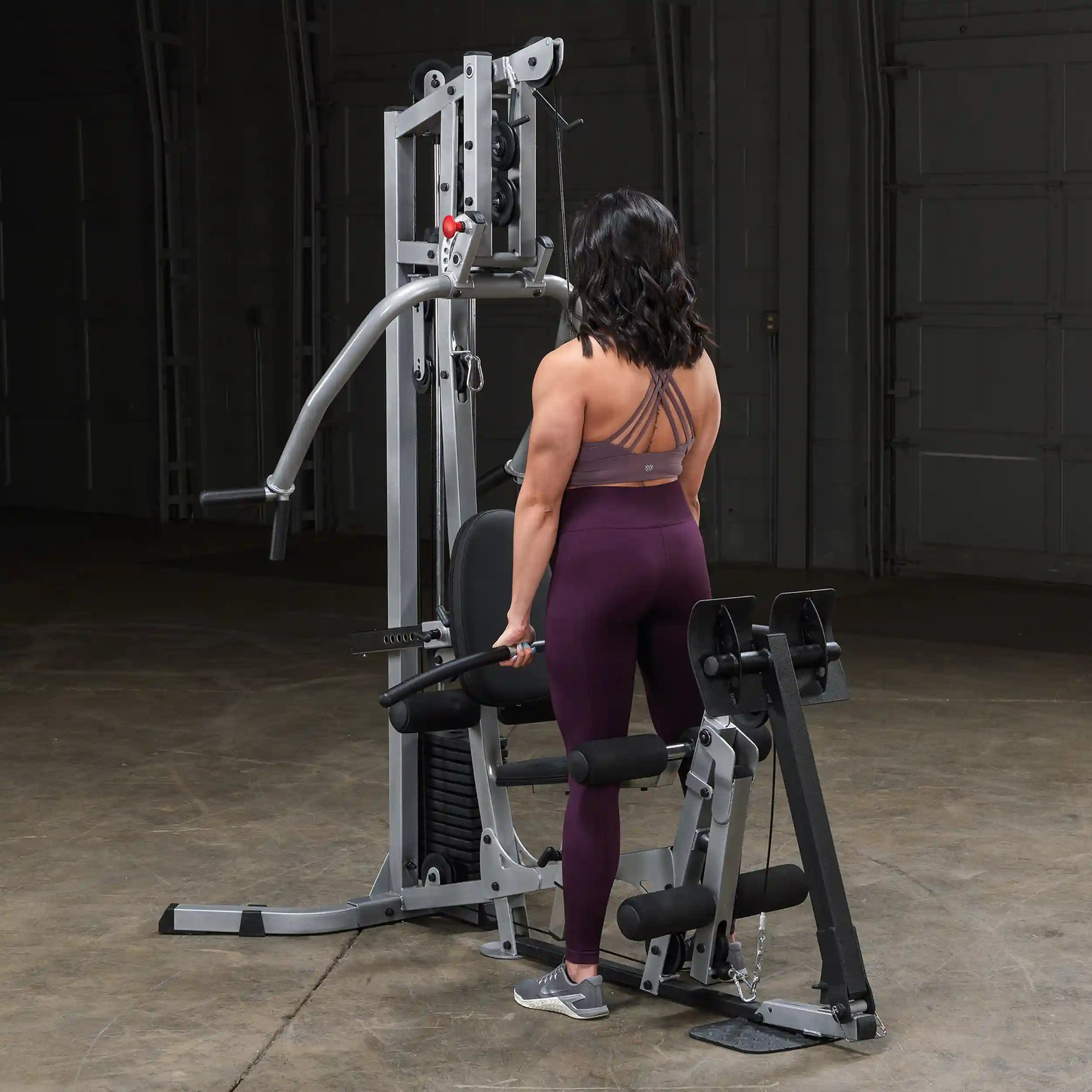 A person with shoulder-length dark hair, wearing a purple sports bra and matching leggings, stands at a gym machine, preparing to perform an exercise in a well-lit indoor space.