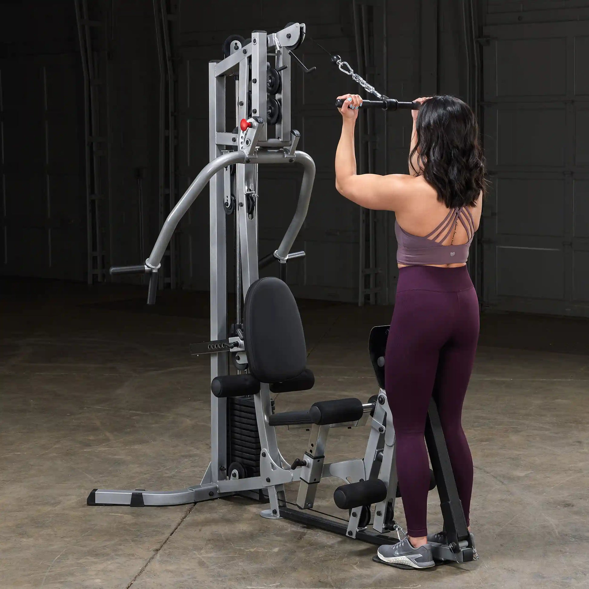 A woman with shoulder-length dark hair, wearing a purple sports bra and high-waisted leggings, performs an exercise on a multi-gym machine, focusing on her upper body workout.