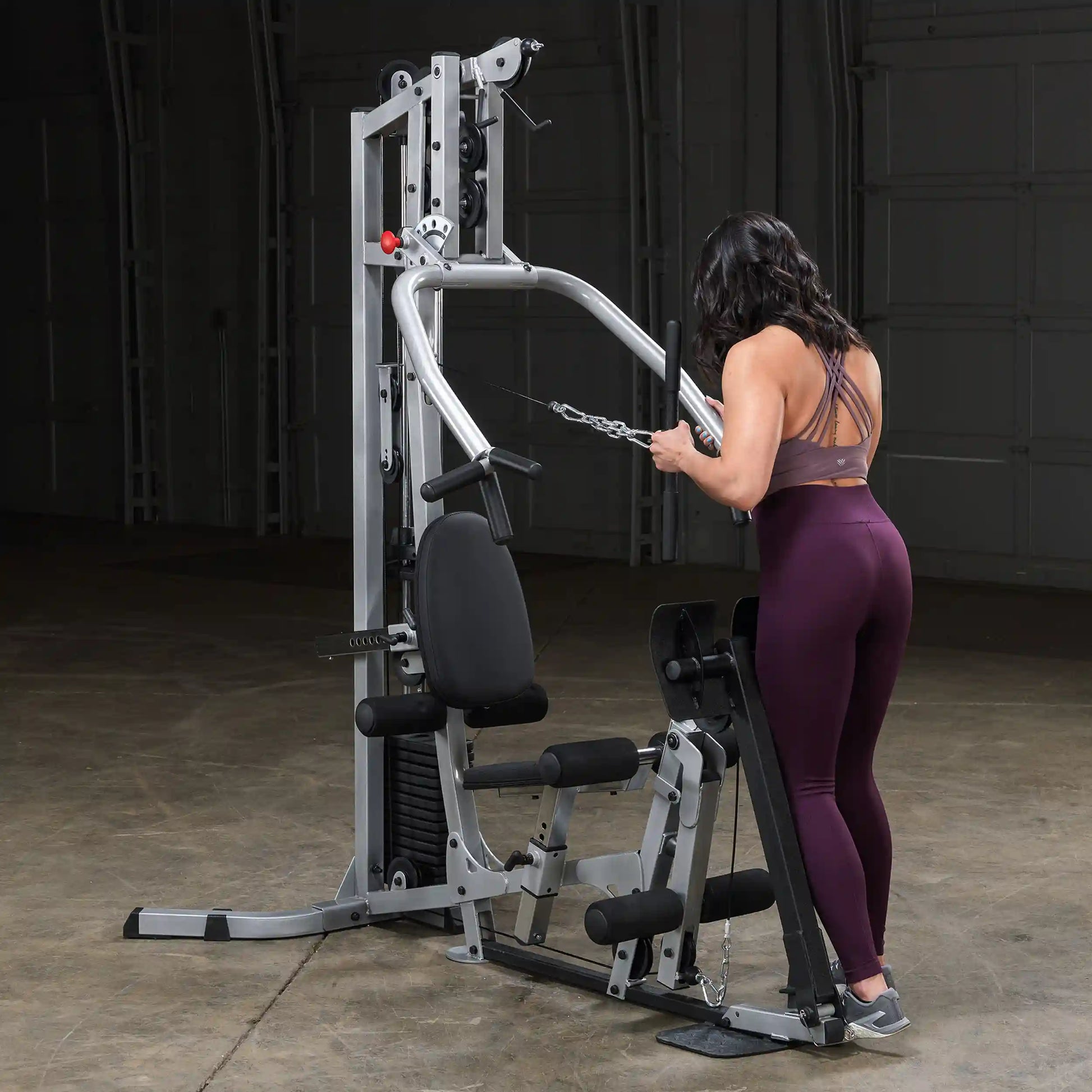 A woman in athletic wear is using a multi-gym machine for strength training, focusing on her back muscles. The gym equipment is positioned in a spacious, dimly lit area with a smooth floor.