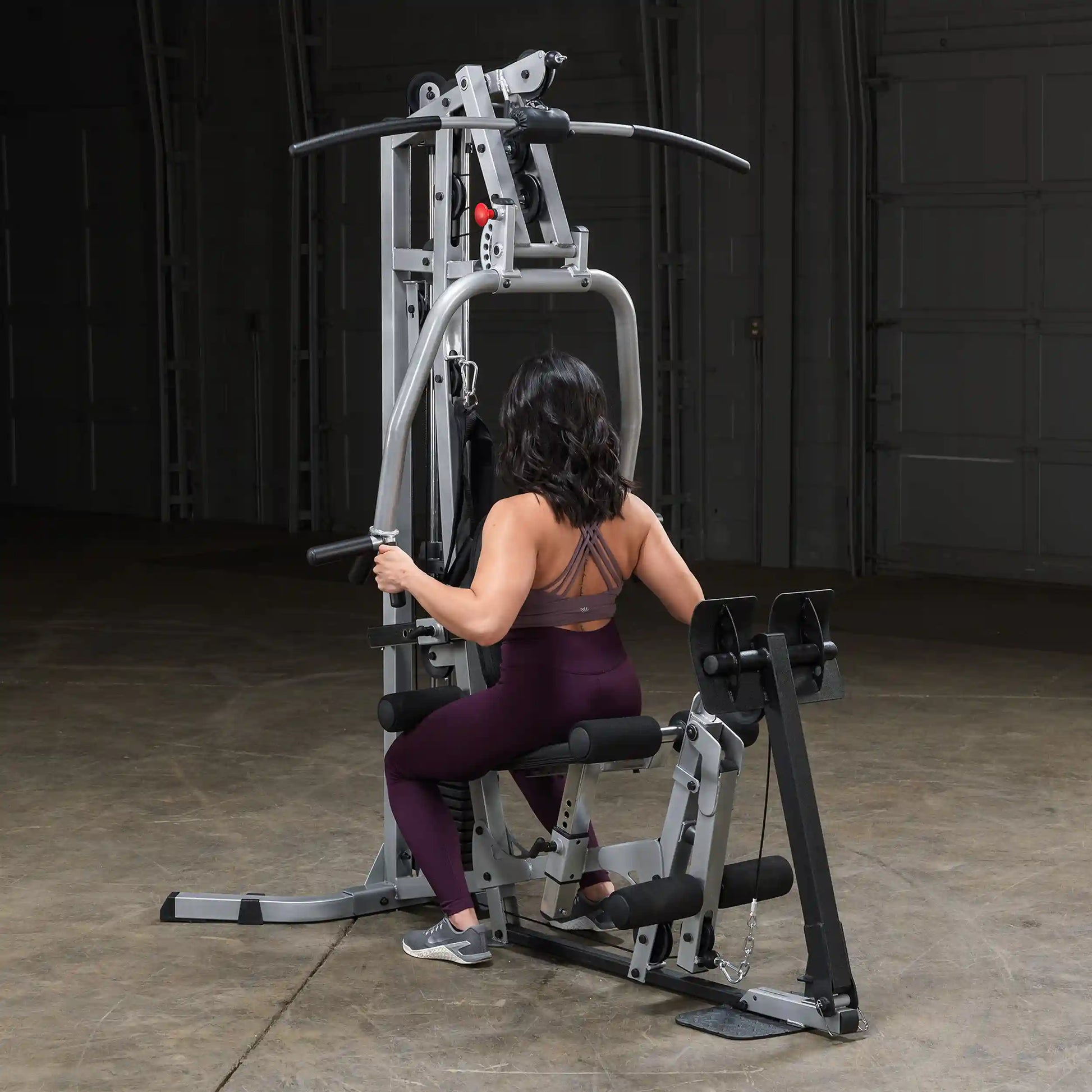 A person with dark, wavy hair is seated on a multi-gym workout machine, engaging in exercise. They are wearing a brown sports bra and maroon leggings, with a focus on strength training in a spacious, well-lit gym environment.