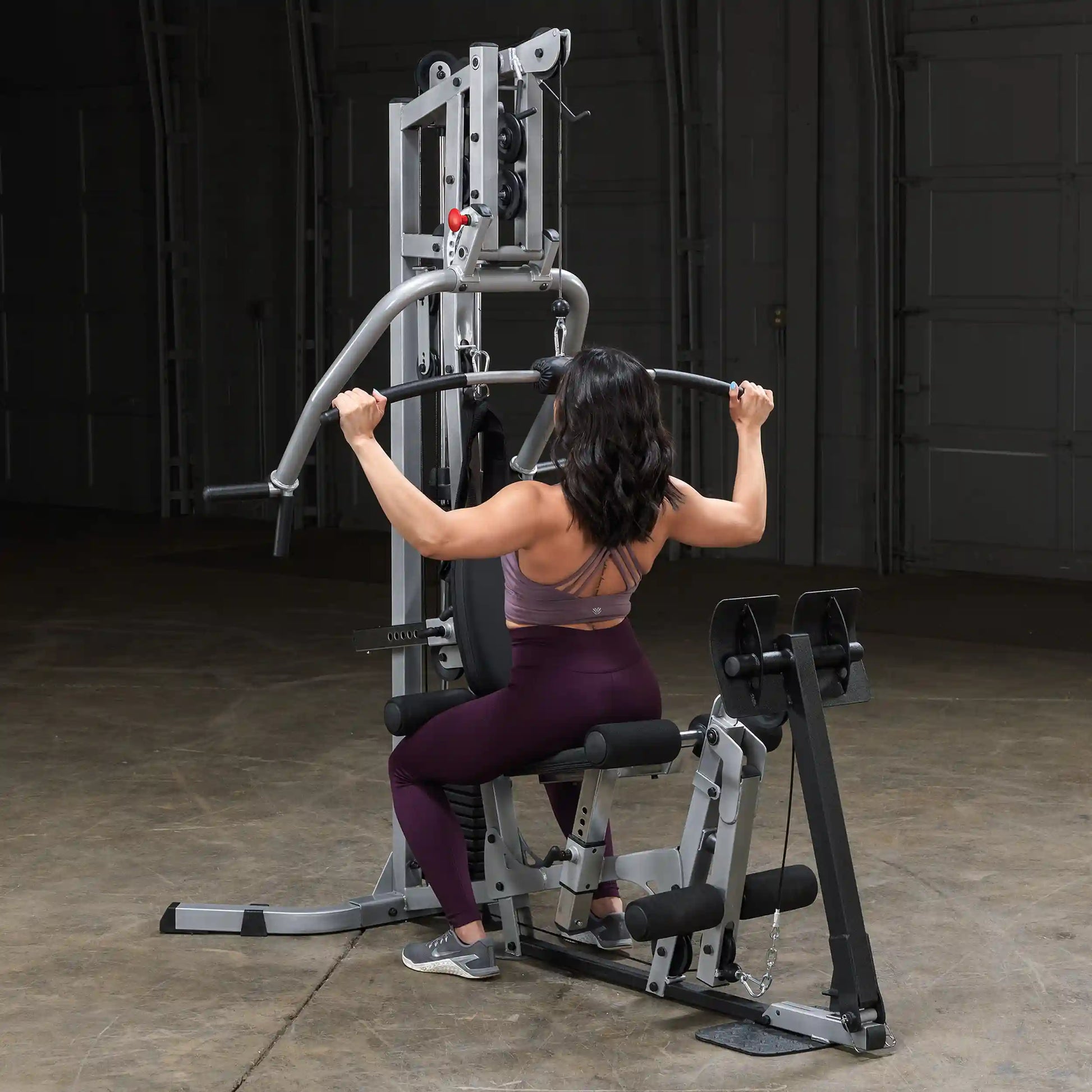 A person with long dark hair is using a multi-gym machine for strength training. They are seated and gripping a bar, with weights visible on the machine. The setting is a spacious indoor gym environment.