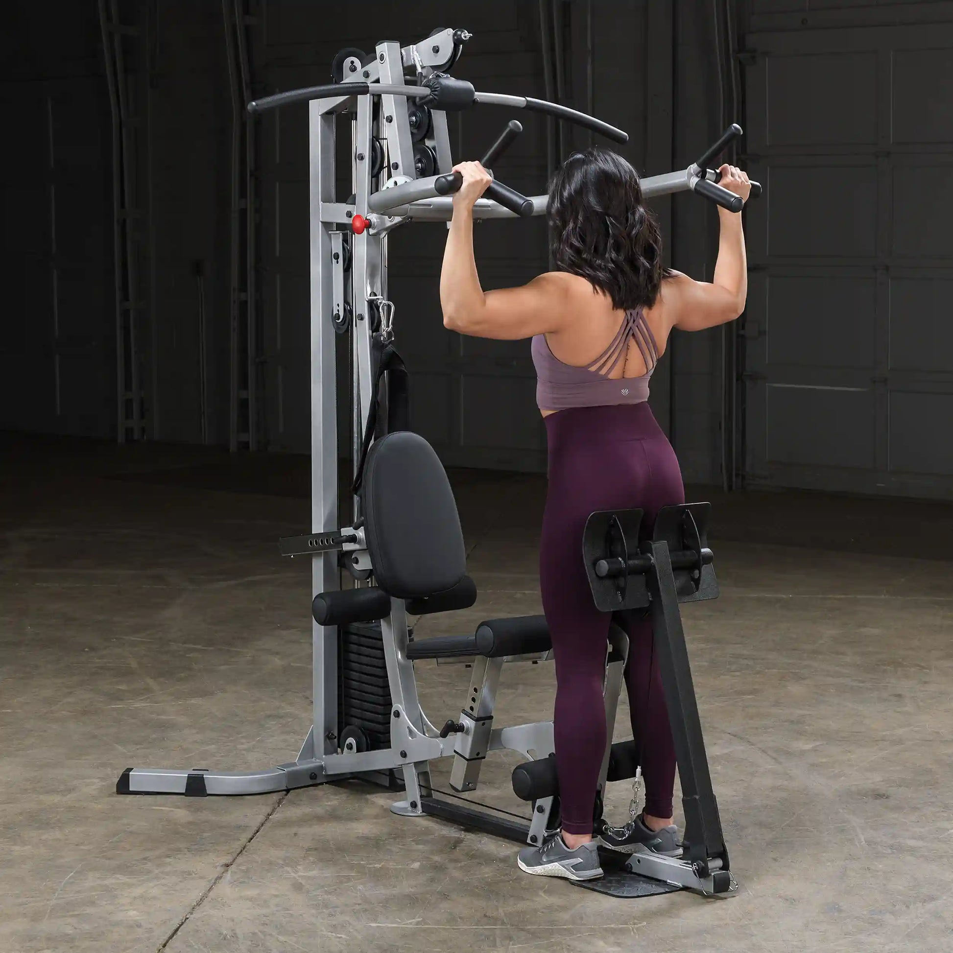 A woman performing a pull-up on a multi-gym machine in a gym setting, wearing a purple sports bra and maroon leggings, with her back facing the camera.