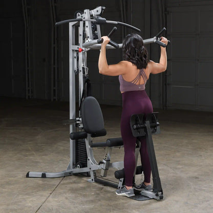 A woman performing a pull-up on a multi-gym machine in a gym setting, wearing a purple sports bra and maroon leggings, with her back facing the camera.