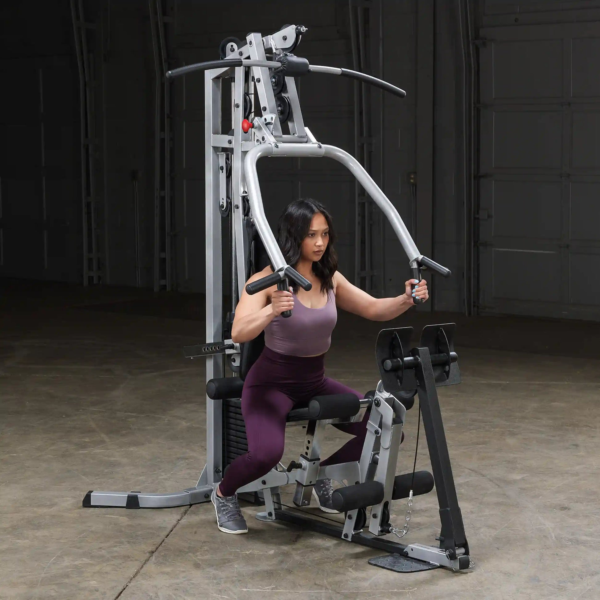 A woman in athletic wear performs an exercise on a weight machine in a gym setting. The machine has multiple handles and weights, and the background is dimly lit.