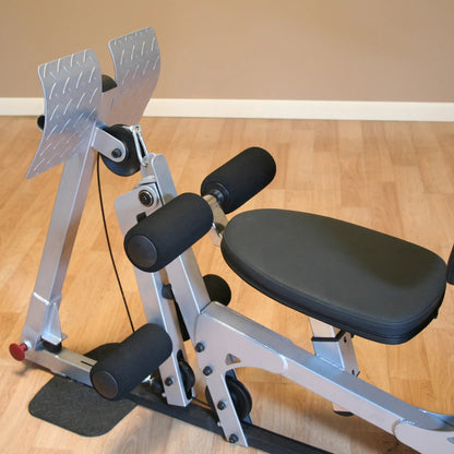 Close-up of a silver exercise machine with padded seat and rollers, designed for leg workouts, set on a wooden floor.