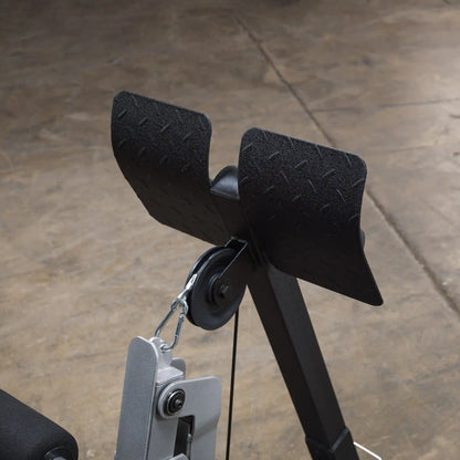 Close-up view of a gym equipment attachment featuring two padded supports and a pulley system, set against a textured floor background.