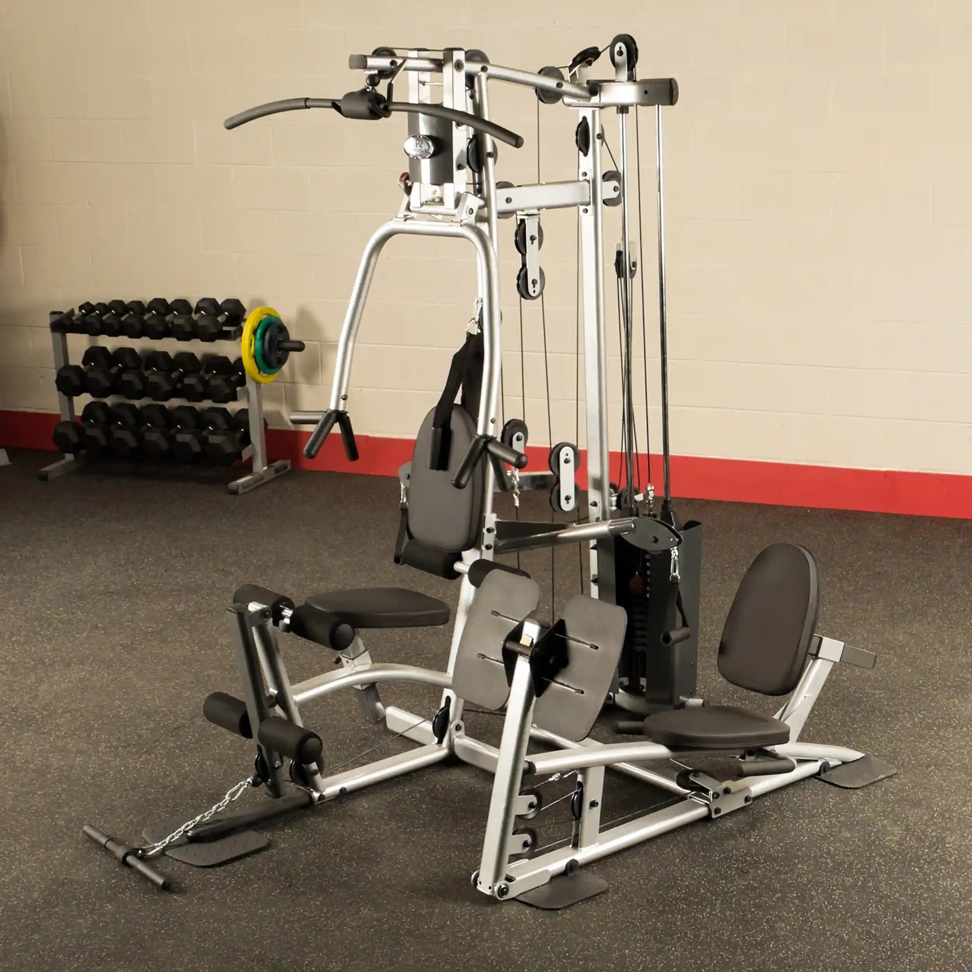 A multi-station home gym equipment featuring various workout stations, including a lat pull-down, leg extension, and cable pulley system, with a rack of dumbbells in the background.