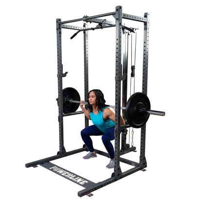 A woman performing a squat with a barbell on her shoulders inside a squat rack, wearing a blue tank top and leggings, with weight plates on the barbell.