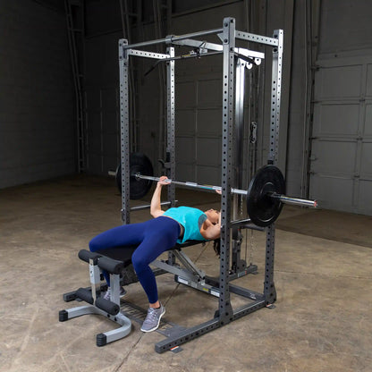 A woman performing a bench press exercise on a weightlifting bench, using a barbell with weights, inside a gym setting. She is wearing a blue tank top and dark leggings, with her hair tied back. The gym has a gray floor and equipment in the background.