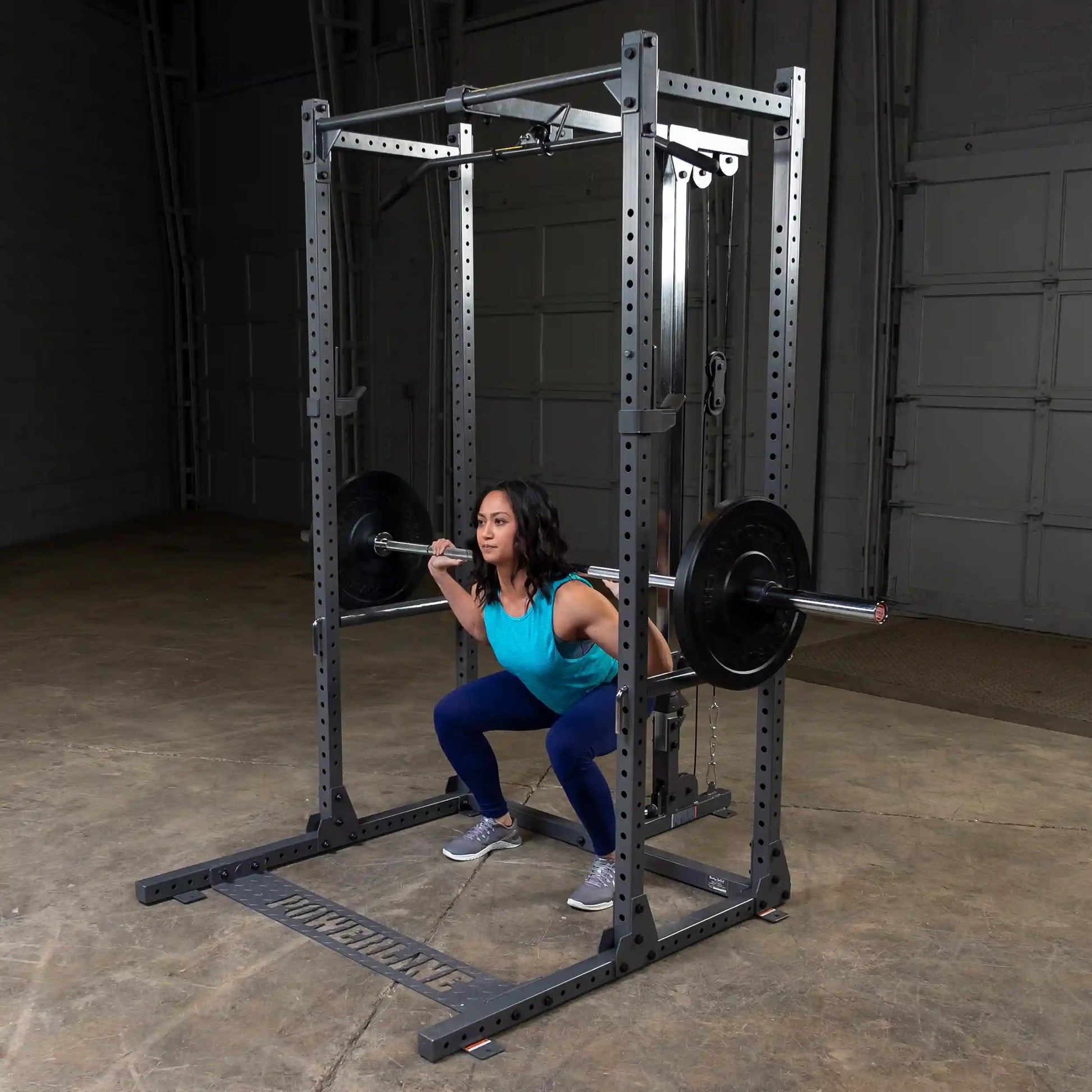 A woman performing a squat with a barbell on her shoulders inside a gym, using a squat rack. She is wearing a turquoise tank top and blue leggings, and the gym has a concrete floor and a gray backdrop.