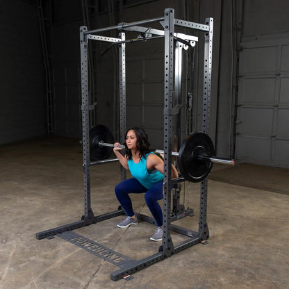 A woman performing a squat with a barbell on her shoulders inside a gym, using a squat rack. She is wearing a turquoise tank top and blue leggings, and the gym has a concrete floor and a gray backdrop.