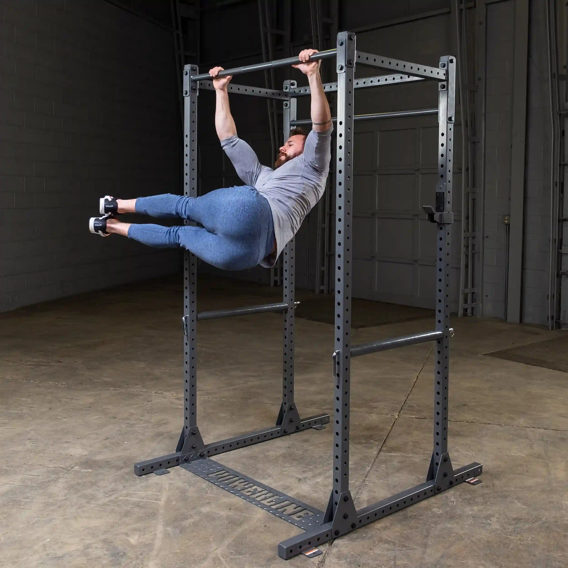 A man performing a hanging leg raise on a pull-up bar in a spacious gym environment, wearing a gray long-sleeve shirt and blue leggings.