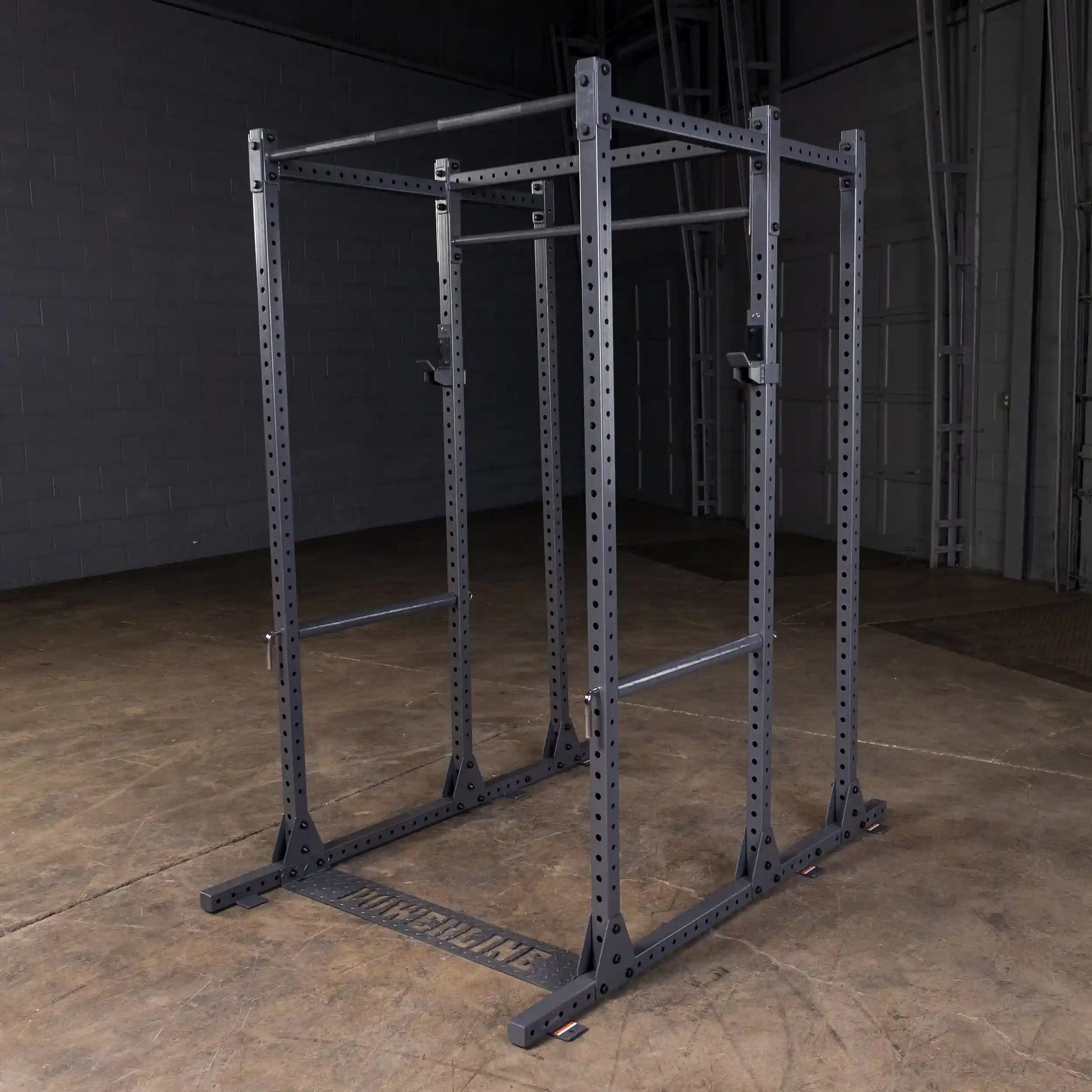 A black power rack with adjustable bars, positioned in an empty gym space with a concrete floor and gray walls.