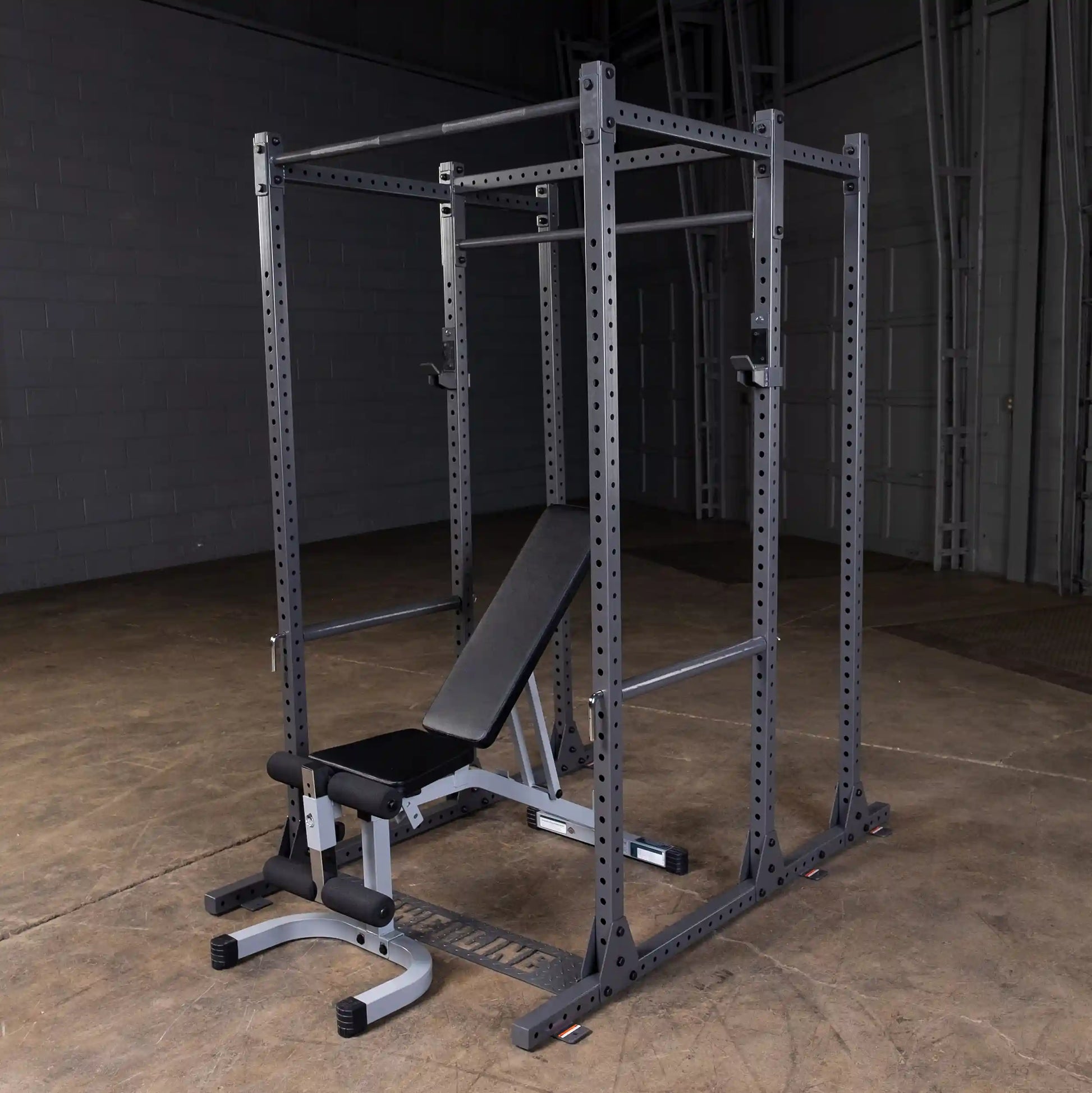 A sturdy gray power rack with an adjustable bench in a spacious gym environment, featuring a concrete floor and dim lighting.