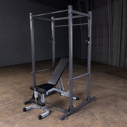 A black and silver squat rack with an adjustable bench positioned in front, set against a concrete floor and wall in a dimly lit gym environment.