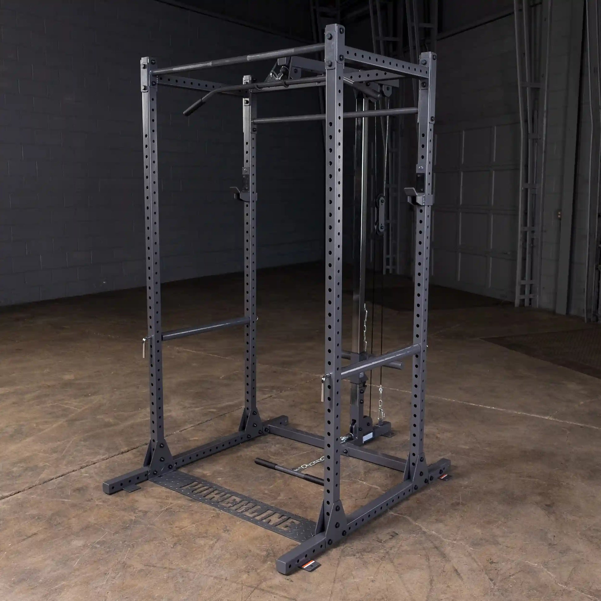 A black power rack with adjustable pull-up bars and cable attachments, set on a wooden floor in a gym environment.