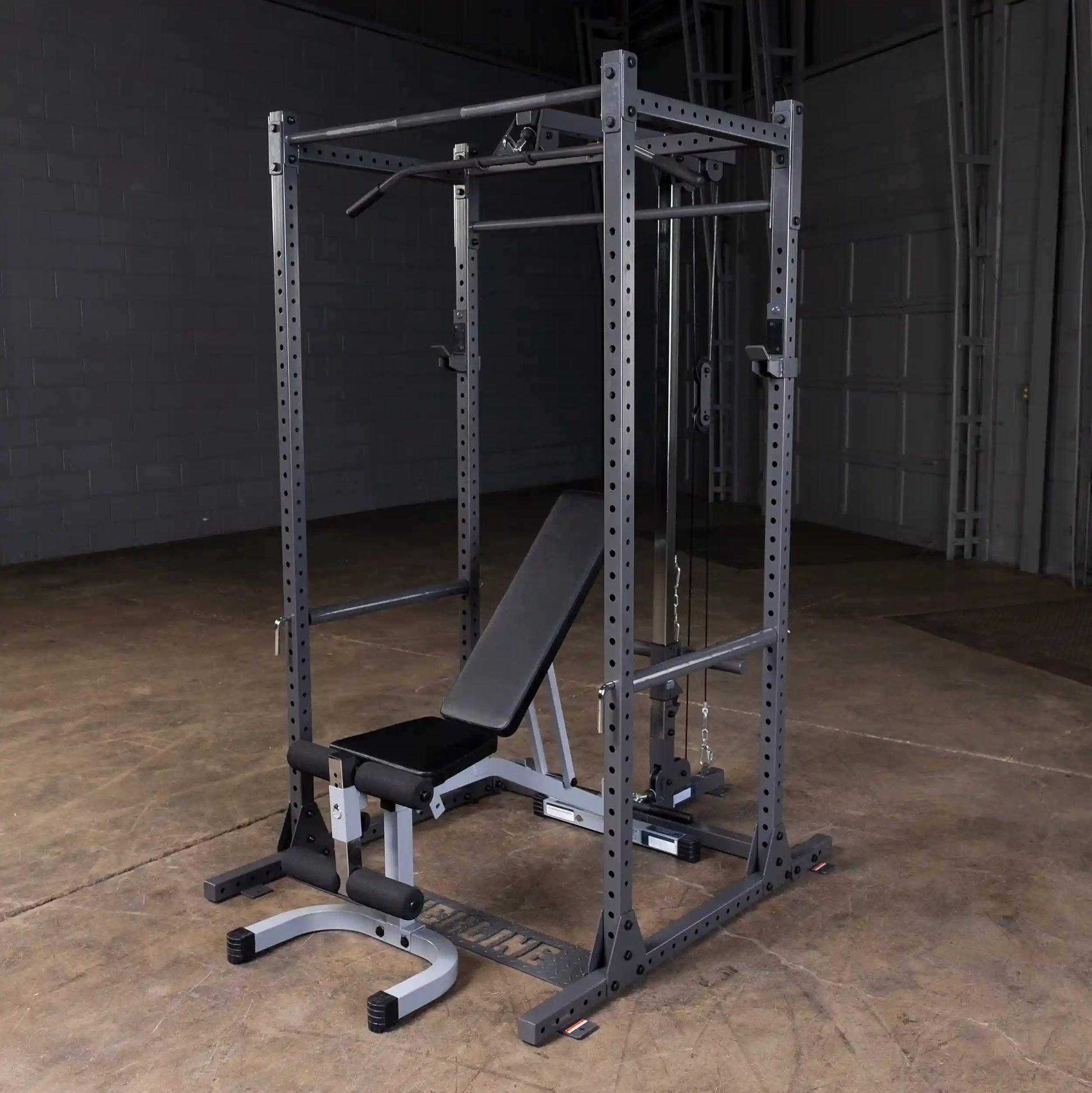 A black and gray multi-functional gym rack with a bench, pull-up bar, and cable attachments, set in a spacious indoor area with a concrete floor.