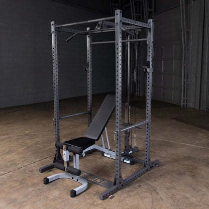 A black and gray multi-functional gym rack with a bench, pull-up bar, and cable attachments, set in a spacious indoor area with a concrete floor.