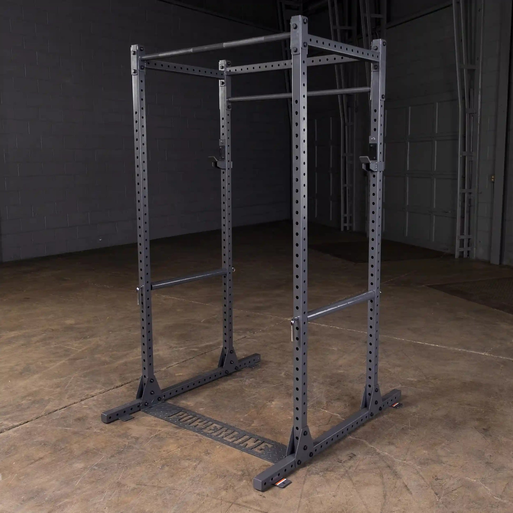 A black power rack with adjustable pull-up bars and a sturdy base, set against a gray wall and wooden floor, designed for strength training and workouts.