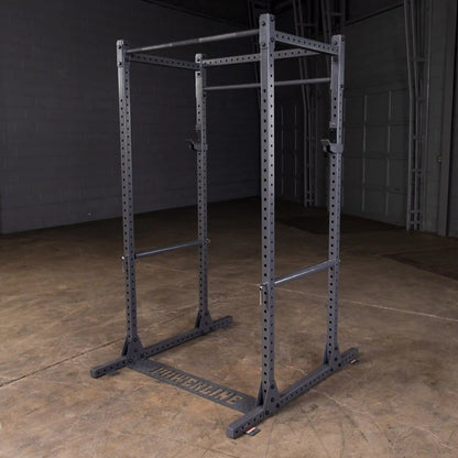 A black power rack with adjustable pull-up bars and a sturdy base, set against a gray wall and wooden floor, designed for strength training and workouts.