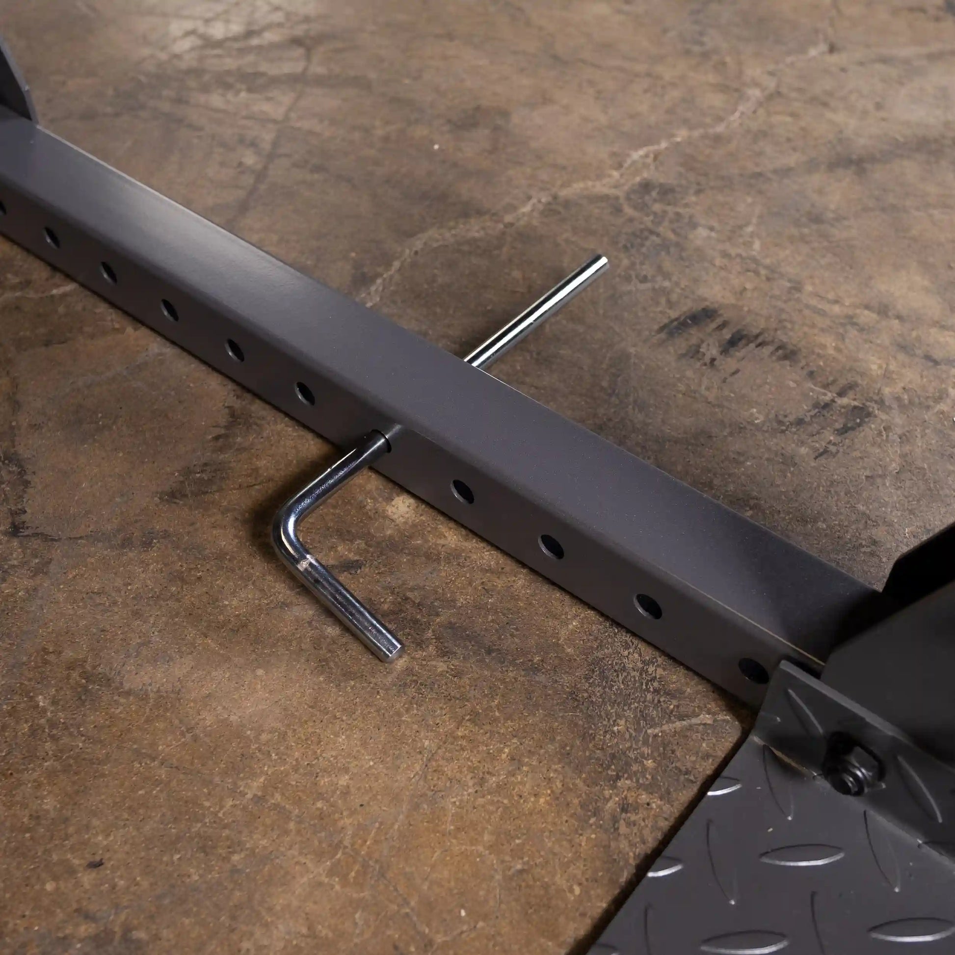 Close-up of a heavy-duty weightlifting bar with adjustment holes and a metallic L-shaped tool resting on a textured surface.