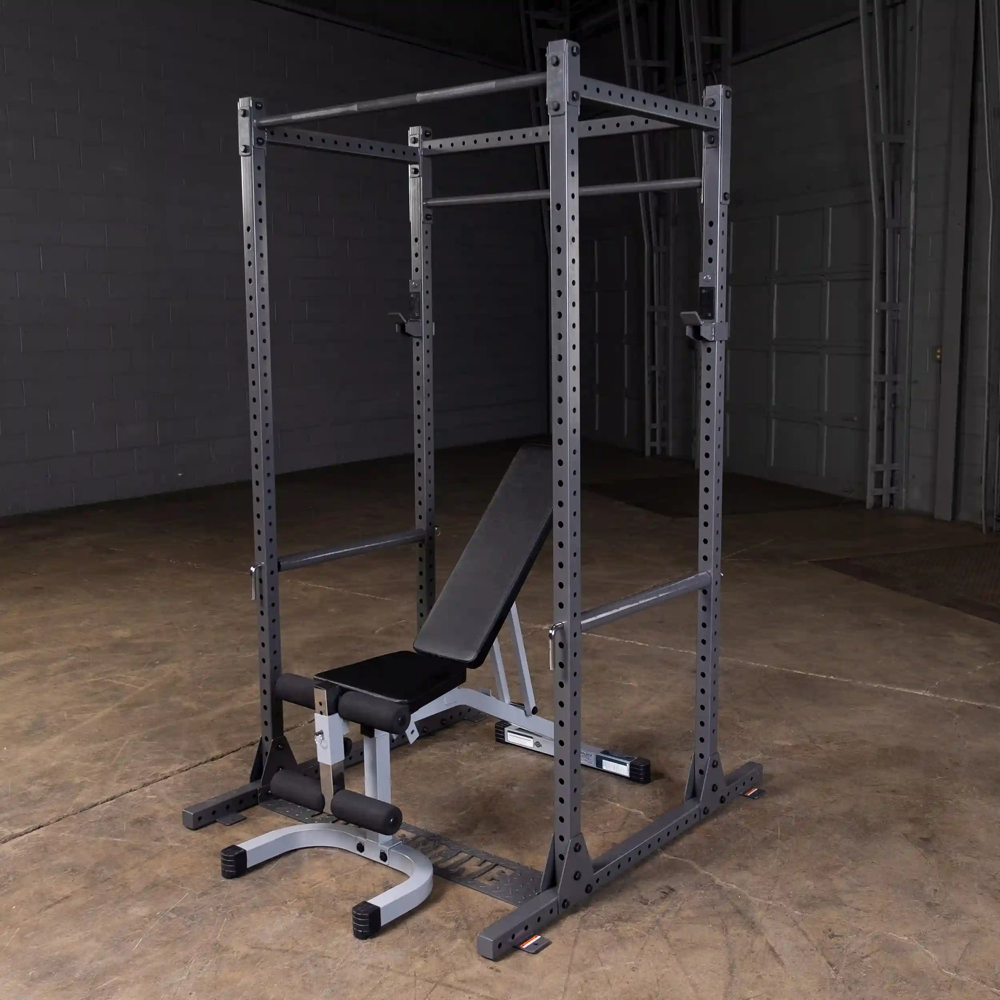 A black squat rack with a bench press setup, featuring adjustable safety bars and a padded bench, situated on a concrete floor in a dimly lit gym environment.