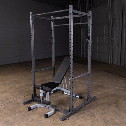 A black squat rack with a bench press setup, featuring adjustable safety bars and a padded bench, situated on a concrete floor in a dimly lit gym environment.