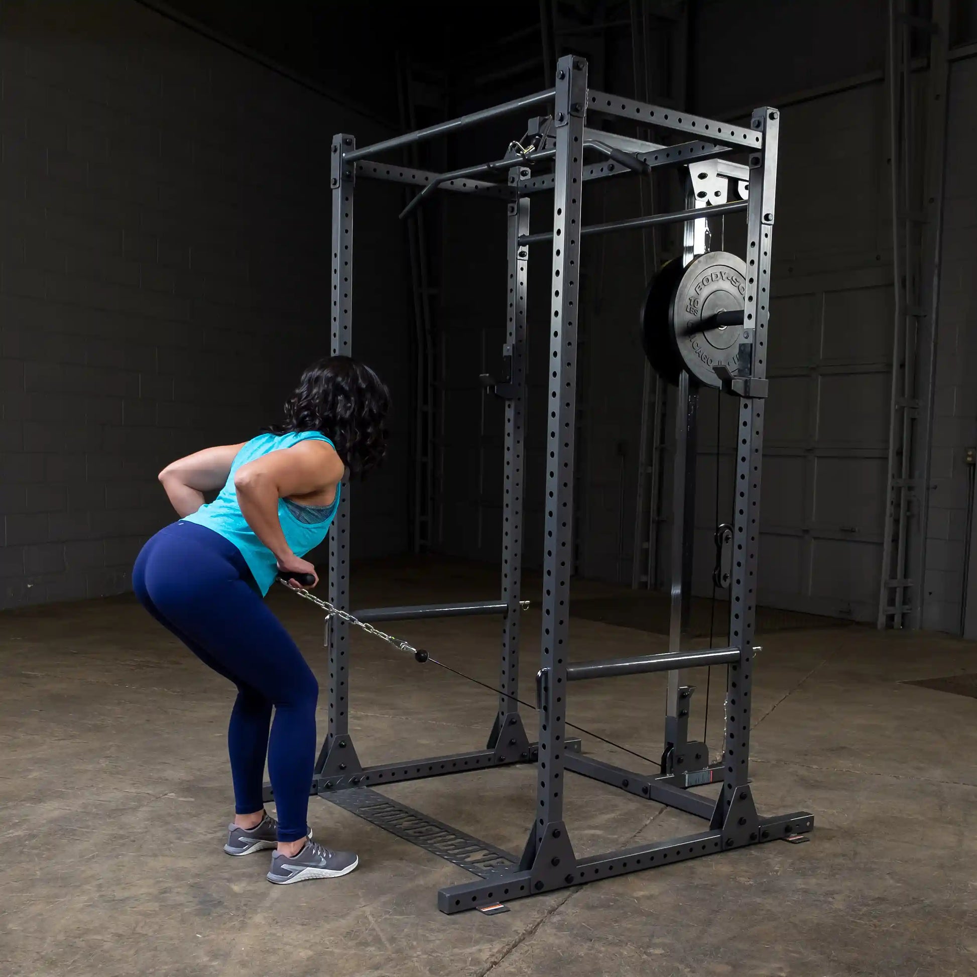 A woman in a turquoise tank top and blue leggings performs a cable row exercise in a gym, using a cable machine with weights attached.
