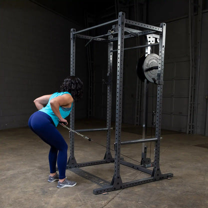 A woman in a turquoise tank top and blue leggings performs a cable row exercise in a gym, using a cable machine with weights attached.