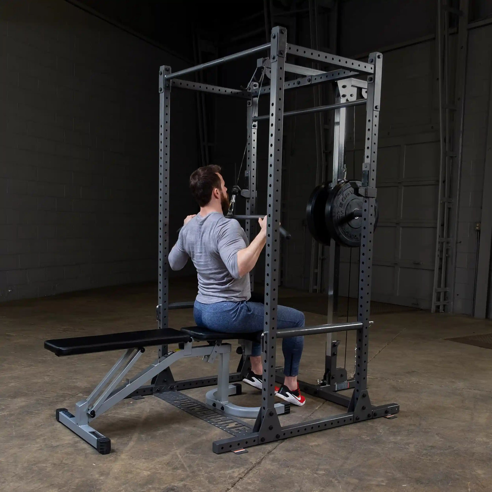 A man performing a seated lat pulldown exercise on a weight machine in a gym setting, wearing a gray long-sleeve shirt and blue pants, with a black weight plate attached to the machine.