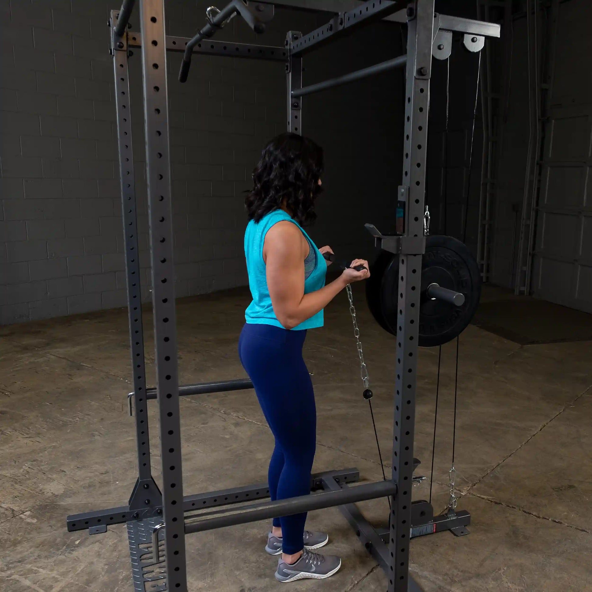 A person in a blue tank top and leggings performs an exercise using a cable machine in a gym setting. The focus is on their form as they pull a chain attached to a weight plate.