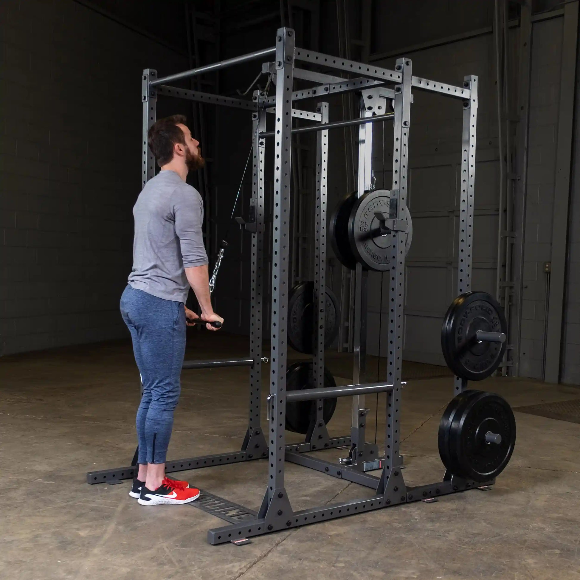A man performing a cable exercise in a gym, using a cable machine with weights. He is wearing a gray long-sleeve shirt, blue workout pants, and red athletic shoes.
