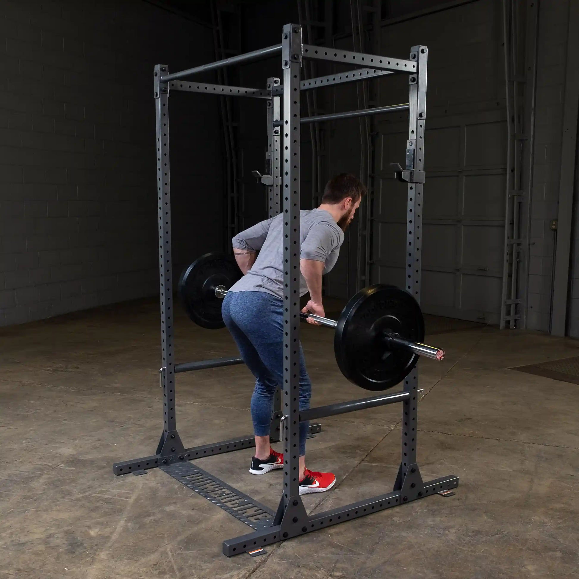 A person performing a deadlift with a barbell in a gym setting. The individual is wearing a gray long-sleeve shirt and blue leggings, with red and white athletic shoes. The barbell is loaded with black weight plates, and the gym has a concrete floor and a rack structure in the background.