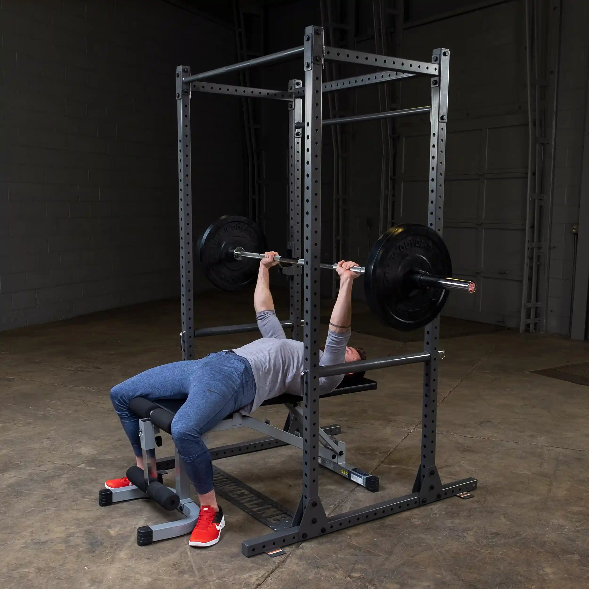 A person performing a bench press exercise with a barbell on a weight bench, wearing a gray long-sleeve shirt and blue athletic pants, in a gym setting with a squat rack and weights.