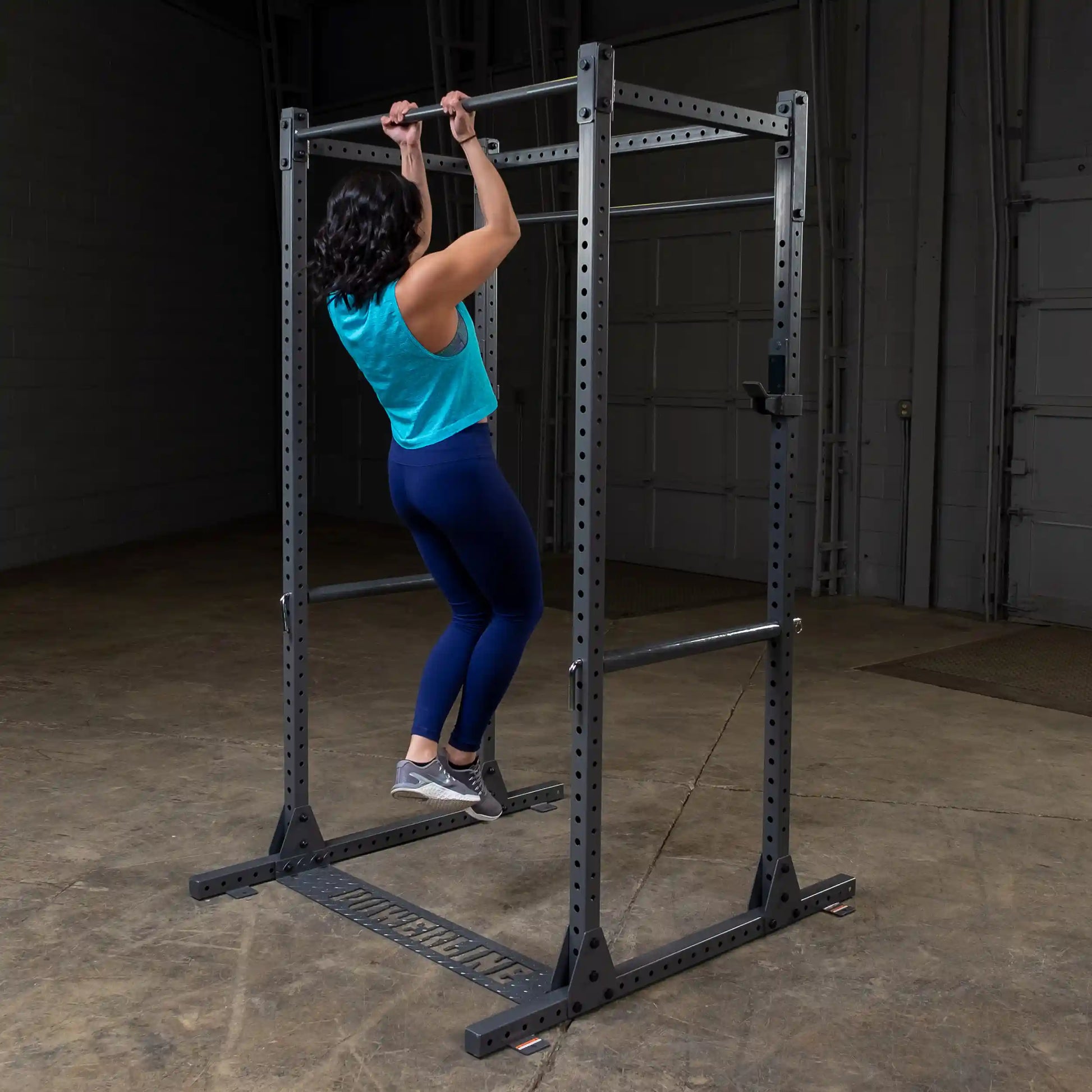 A person performing a pull-up on a metal pull-up bar in a gym setting, wearing a blue tank top and blue leggings, with a focus on strength training.