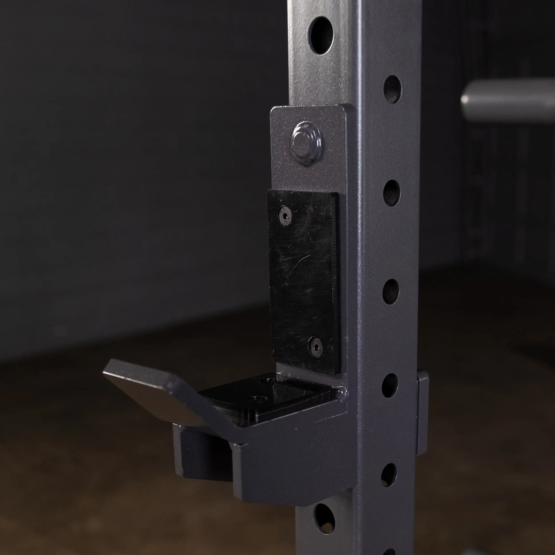 Close-up of a weightlifting rack attachment showing a metal hook on a vertical post with several holes for adjustment, set against a dark background.