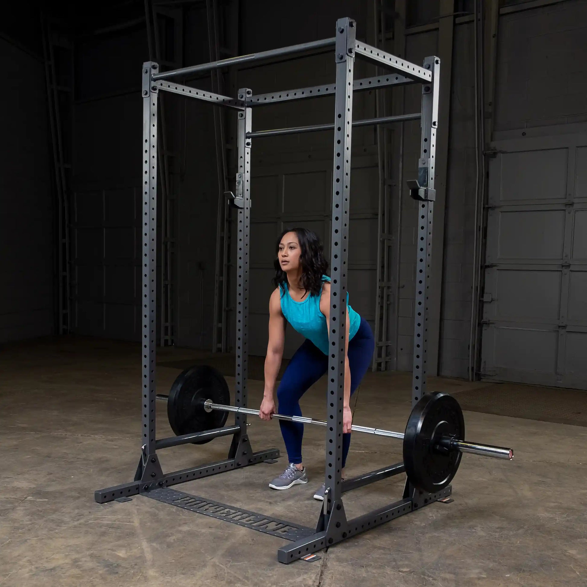 A woman in a turquoise tank top and blue leggings is performing a deadlift in a gym setting, using a barbell with weights on both sides. The gym features a power rack in the background and a concrete floor.
