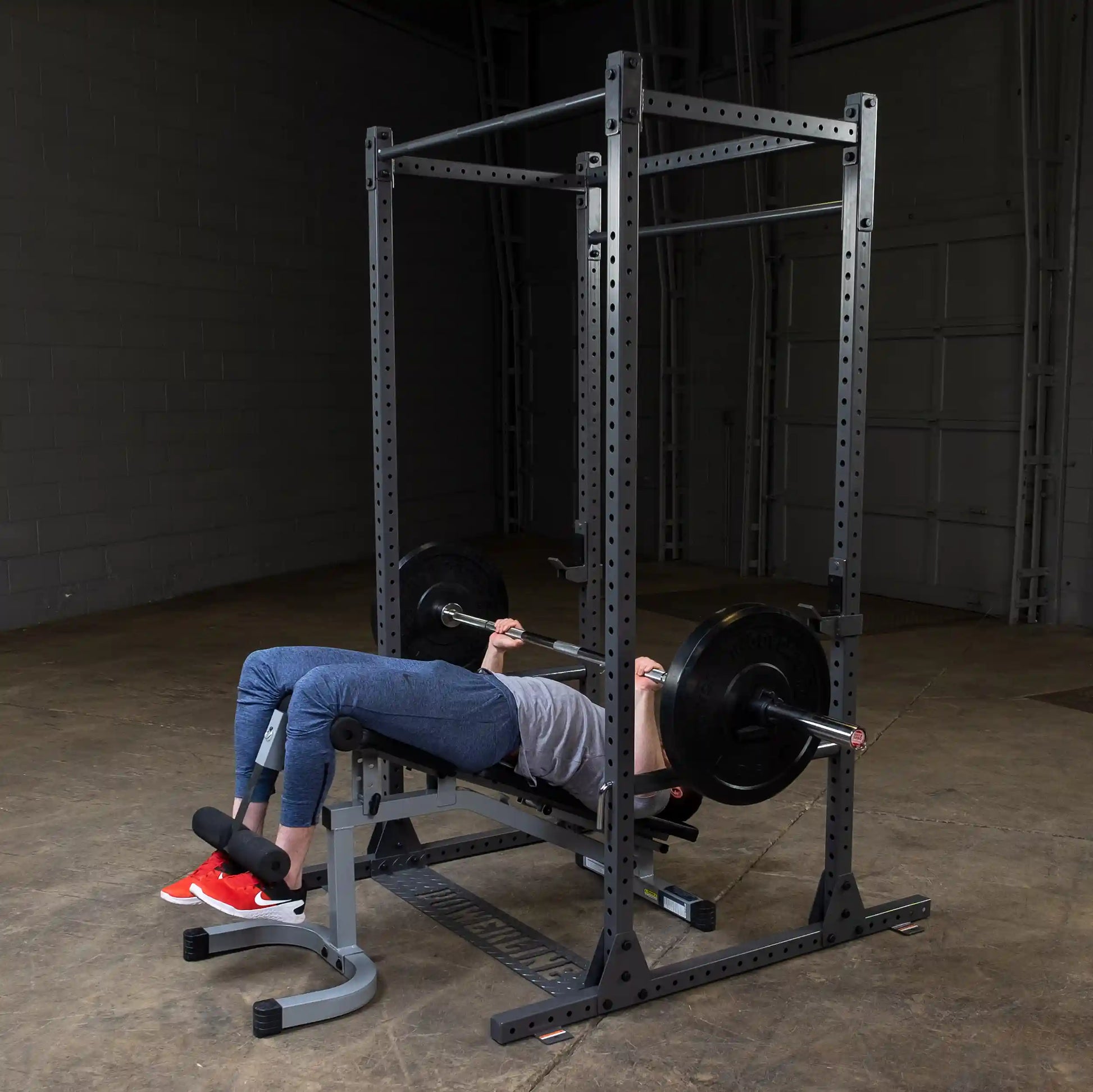 A person performing a bench press exercise on a weightlifting bench with a barbell and weights in a gym setting, wearing gray clothing and red sneakers.