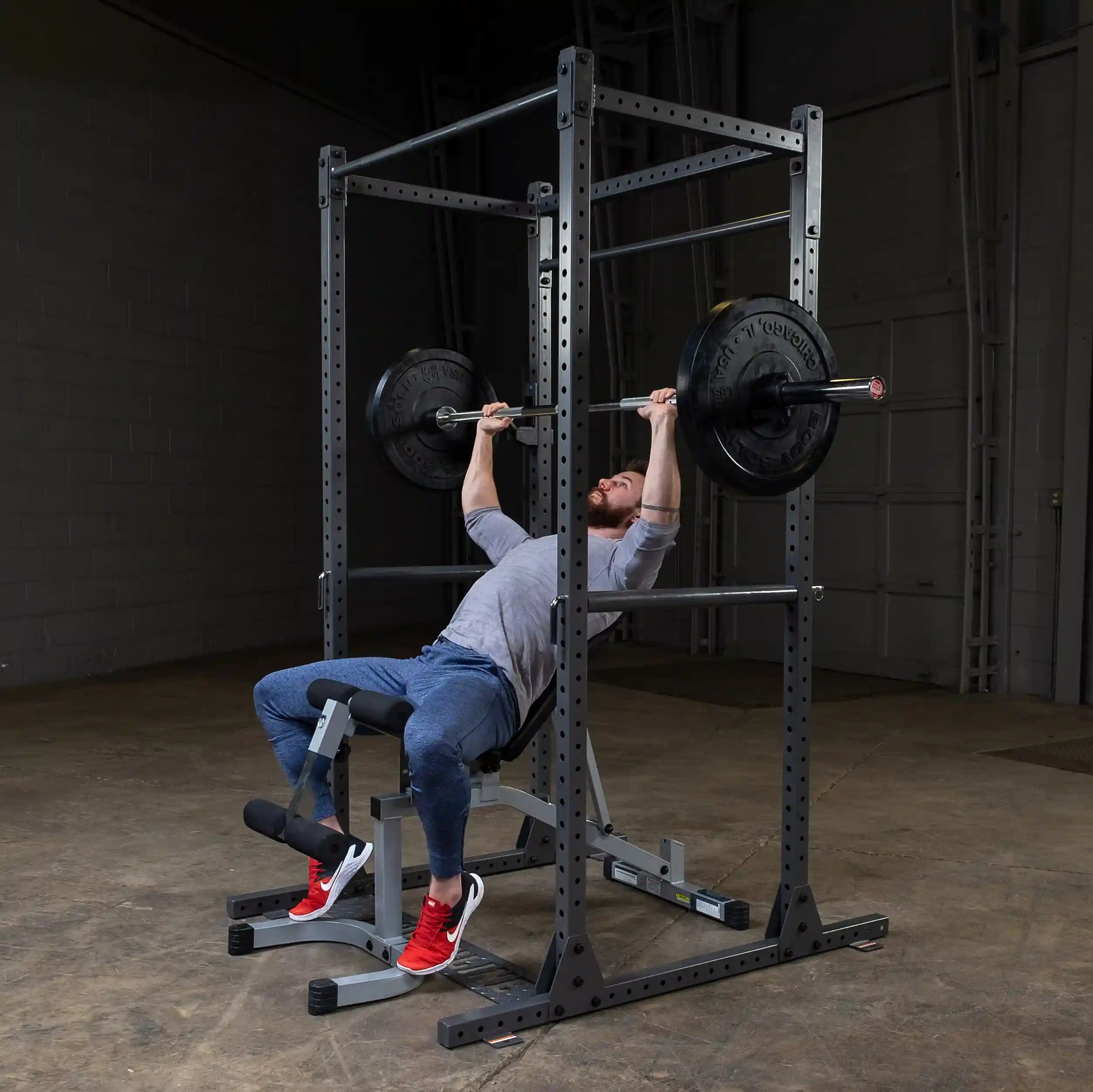 A person performing a bench press exercise with a barbell and weights in a gym setting, wearing a gray shirt, blue pants, and red sneakers, using a squat rack for support.