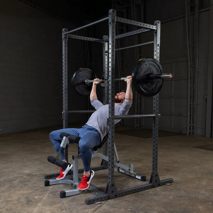 A person performing a bench press exercise with a barbell and weights in a gym setting, wearing a gray shirt, blue pants, and red sneakers, using a squat rack for support.