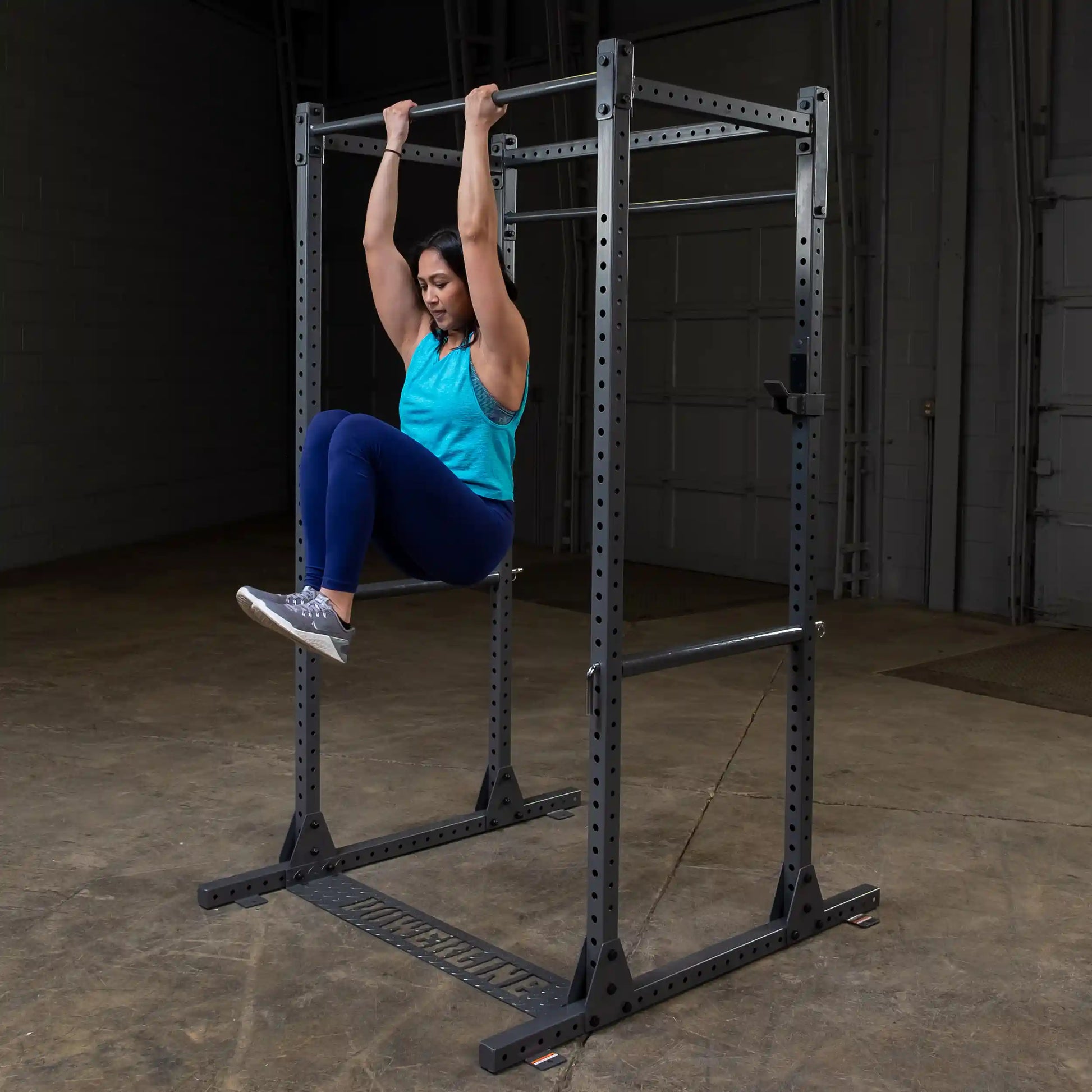 A woman performing a hanging leg raise exercise on a pull-up bar in a gym setting, wearing a blue tank top and blue leggings.
