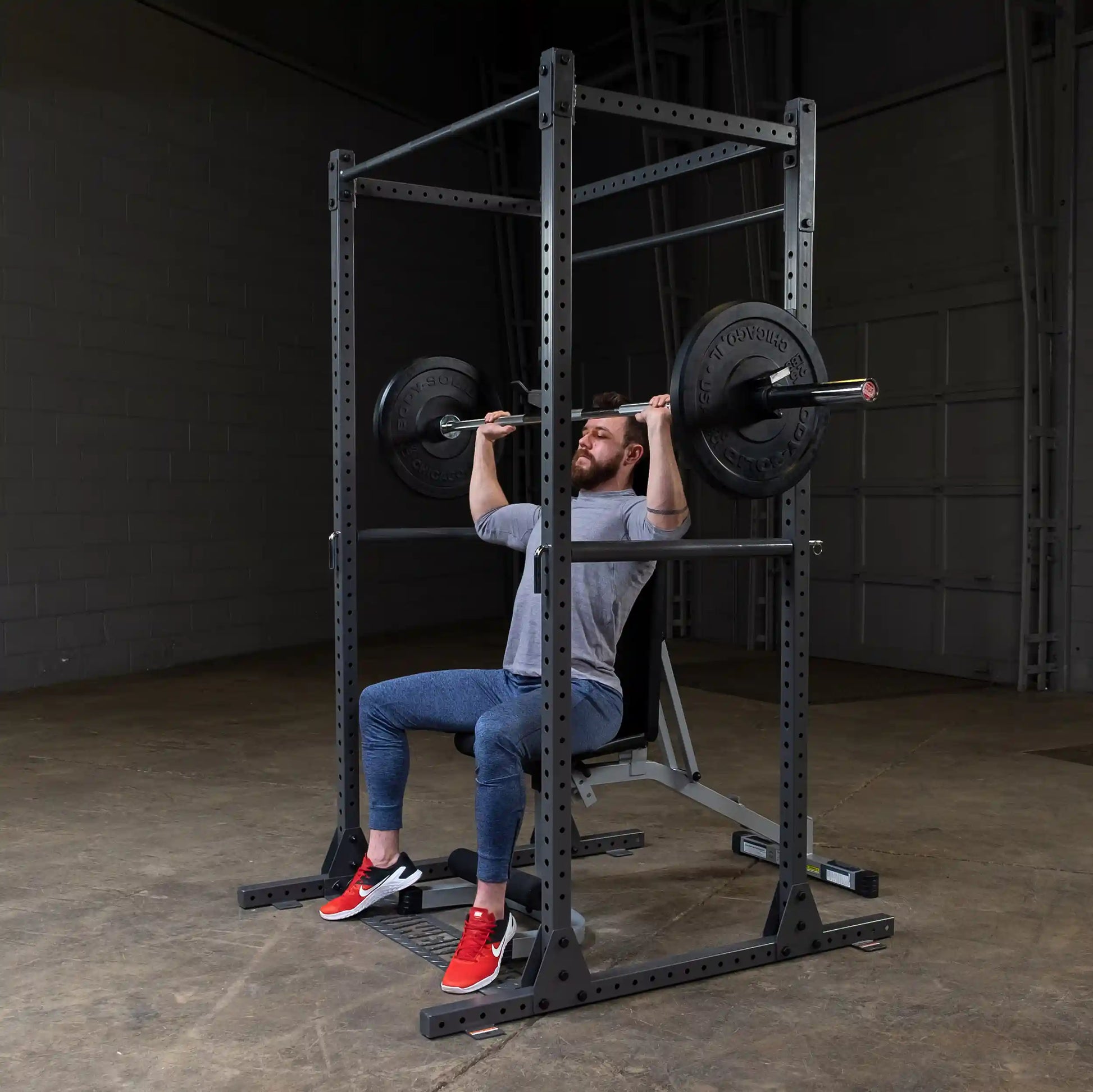 A man performing an overhead press exercise with a barbell on a squat rack, wearing a gray shirt and blue pants, in a gym setting with a concrete floor.