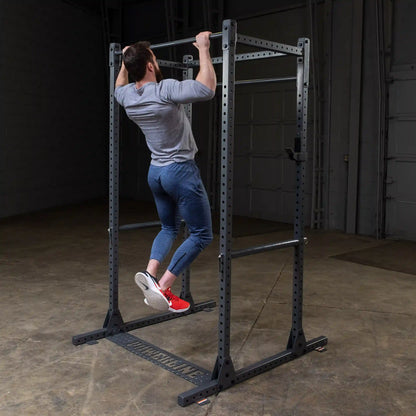 A man performing a pull-up on a black squat rack in a gym setting, wearing a gray long-sleeve shirt and blue athletic pants, with red sneakers.