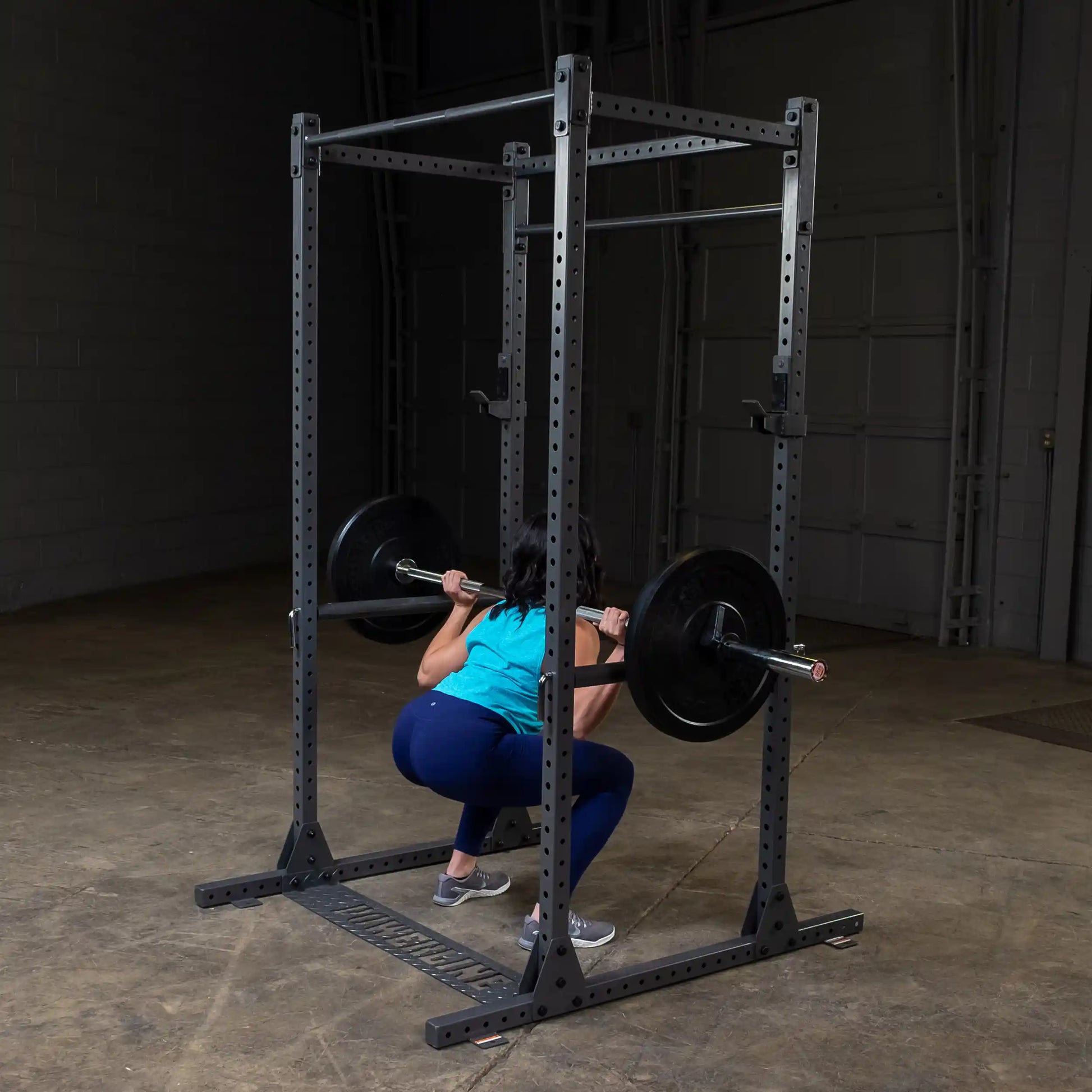A person performing a squat with a barbell on their shoulders in a gym setting, using a squat rack. The individual is wearing a blue tank top and navy leggings, with a focus on proper lifting form.