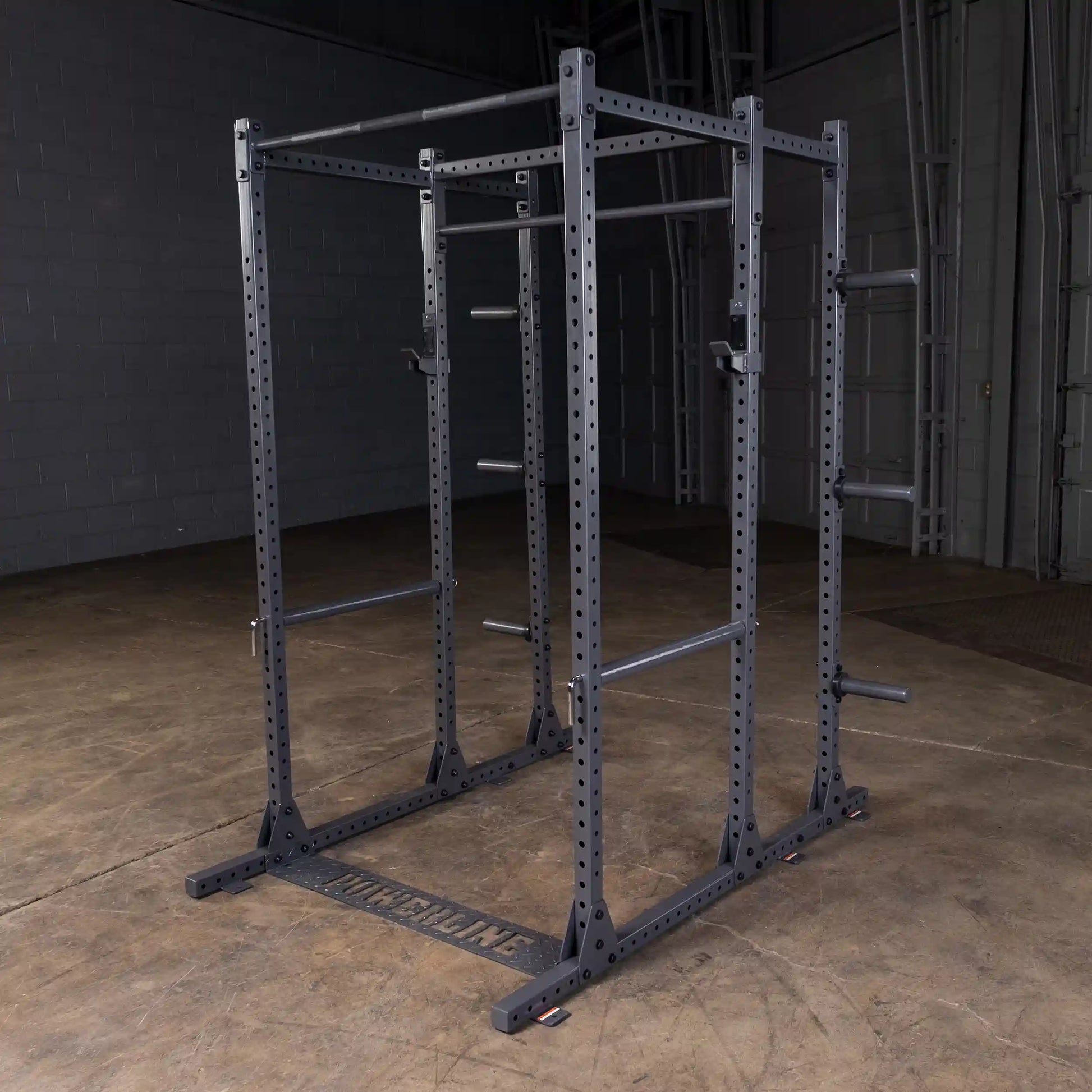 A black power rack with multiple pull-up bars and adjustable safety features, set against a gray wall and concrete floor in a gym environment.
