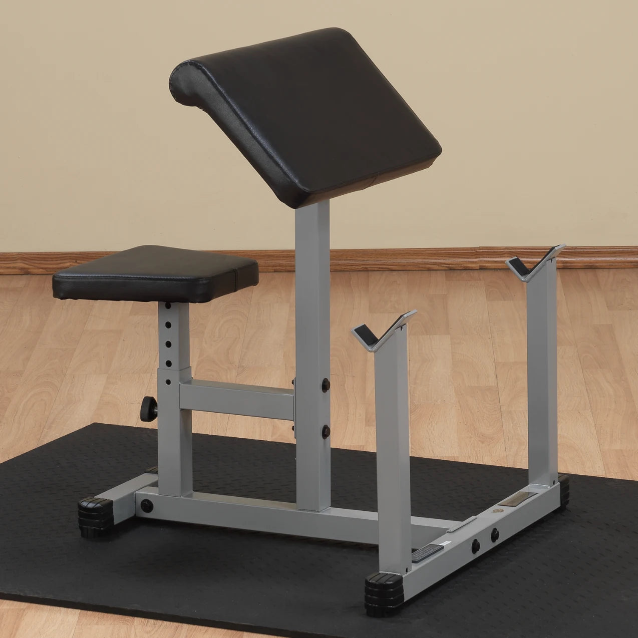 A black and gray weight bench designed for strength training, featuring an adjustable backrest, a padded seat, and two hooks for holding weights, placed on a black mat with a light-colored wall in the background.