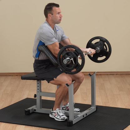 A man performing a seated bicep curl exercise with a barbell, sitting on a weight bench in a gym setting. He is wearing a gray athletic shirt and black shorts, focusing on his workout.
