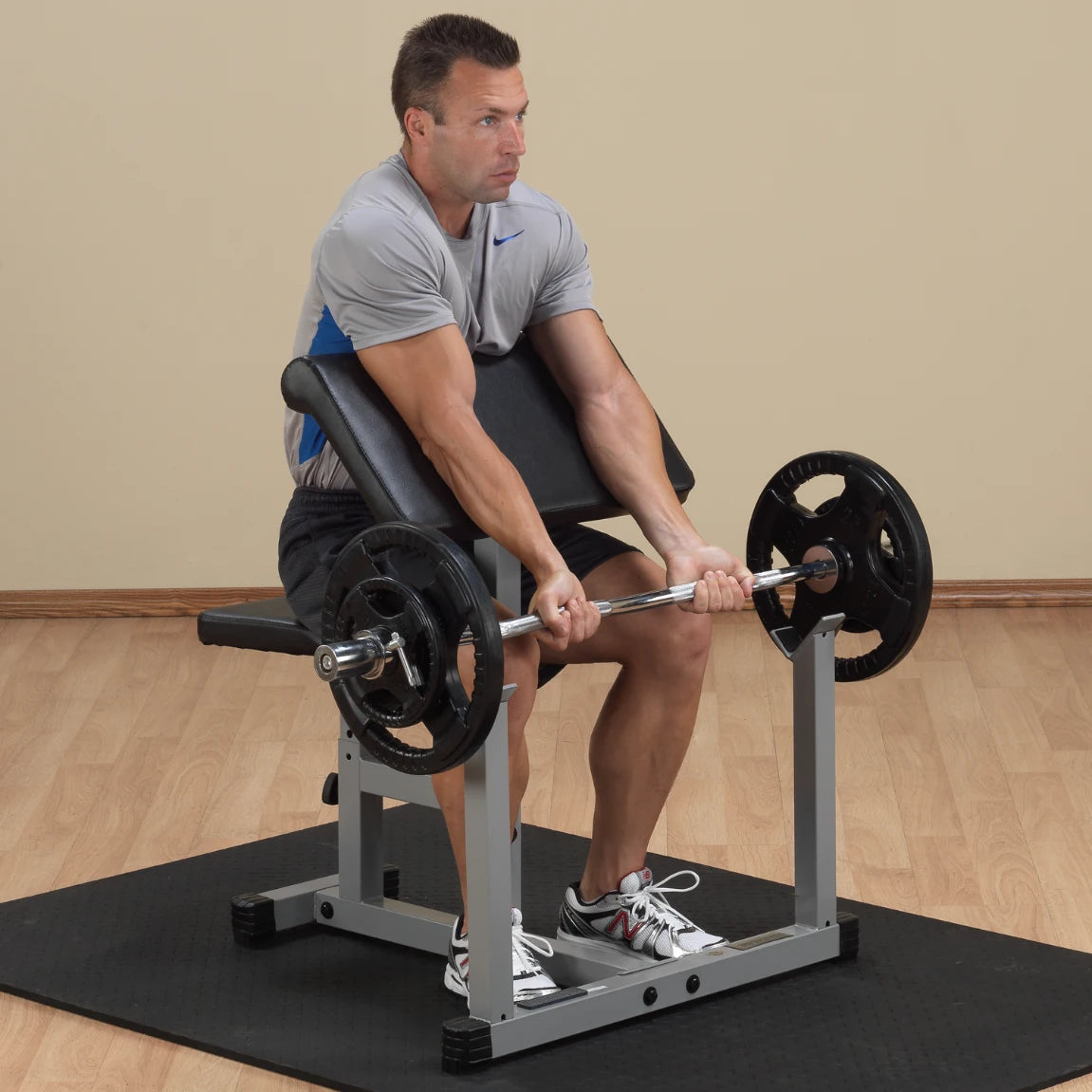 A man performing a seated bicep curl exercise with a barbell on a weight bench in a gym setting.