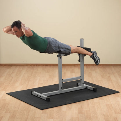A man performing a back extension exercise on a fitness bench, demonstrating proper form with hands behind his head, wearing a green shirt and gray shorts, on a black exercise mat in a light-colored room.