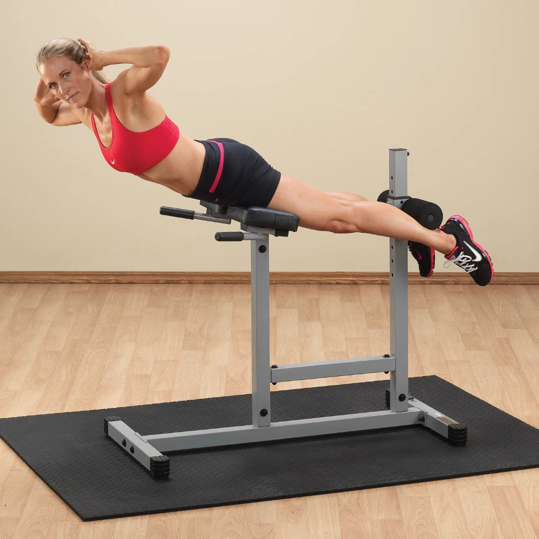 A woman in a red sports bra and black shorts performs an exercise on a decline bench, with her hands behind her head and legs extended, on a rubber mat in a fitness studio.